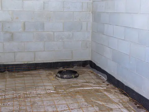 A round black drain basin set in the floor of a bare concrete block basement, with wire mesh lying over wet plastic sheeting around it