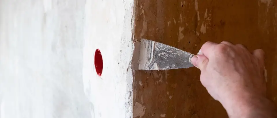 An adult's hand drawing a filling knife loaded with white filler along the edge of a freshly plastered patch on a brown wall