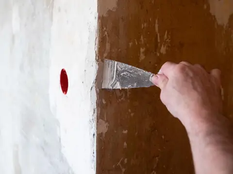 An adult's hand drawing a filling knife loaded with white filler along the edge of a freshly plastered patch on a brown wall