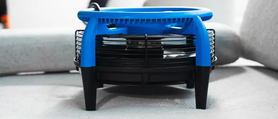 A blue and black air mover fan standing on pale grey upholstery fabric, seen from the side