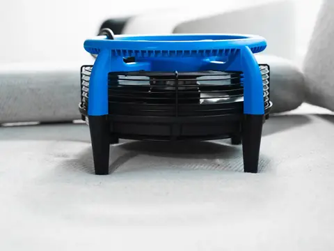 A blue and black air mover fan standing on pale grey upholstery fabric, seen from the side